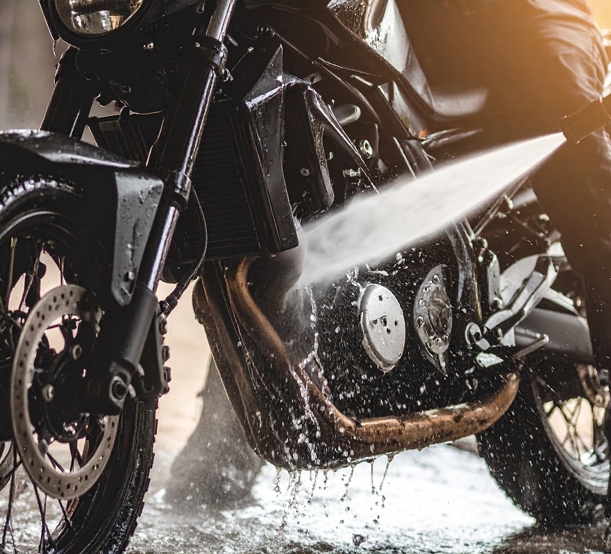 Lavage haute pression d'une moto noire, projetant de l'eau sur le moteur et le pot d'échappement cuivré, avec un effet de lumière.