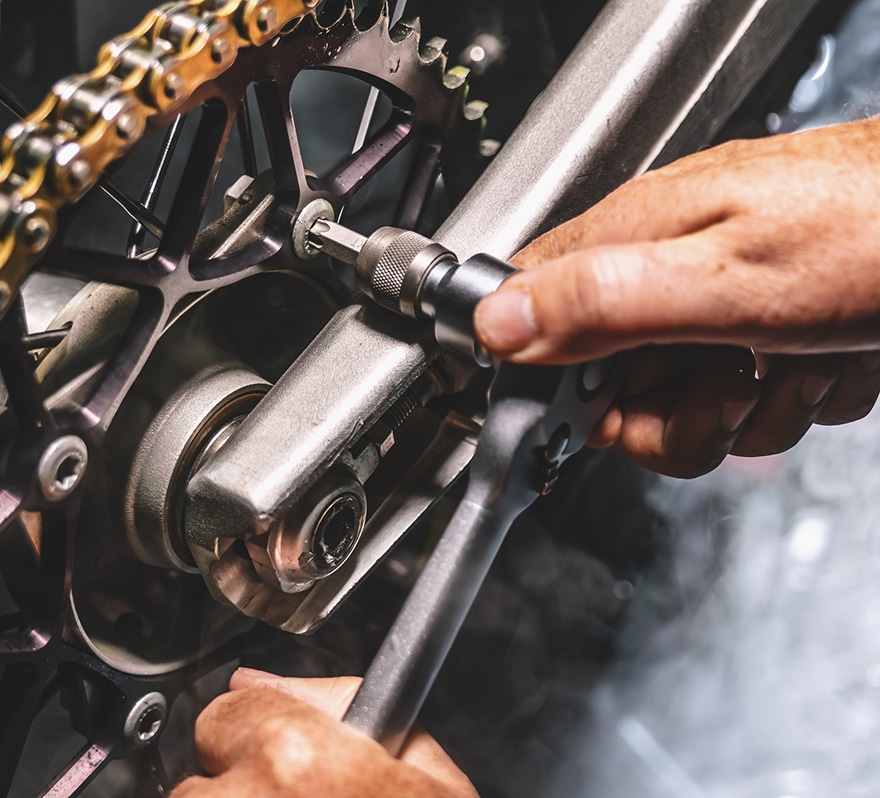 Mécanicien serrant un boulon Torx sur le bras oscillant d'une moto avec une clé à cliquet et une douille, près de la chaîne et du pignon.