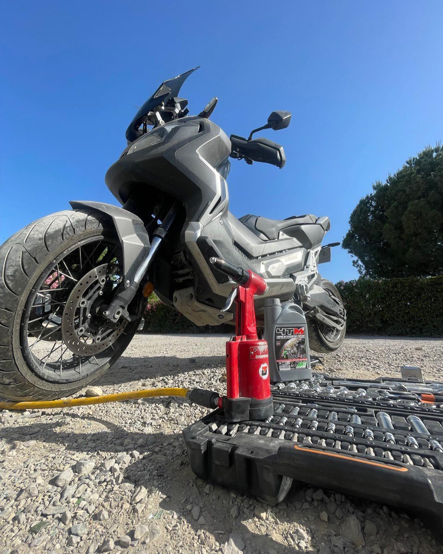 Entretien Honda X-ADV : Outils, Huile HTM et Scooter Moto-scooter Honda X-ADV gris entretenu sur gravier avec clé à chocs rouge, huile moteur HTM 10W40 et jeu de douilles.