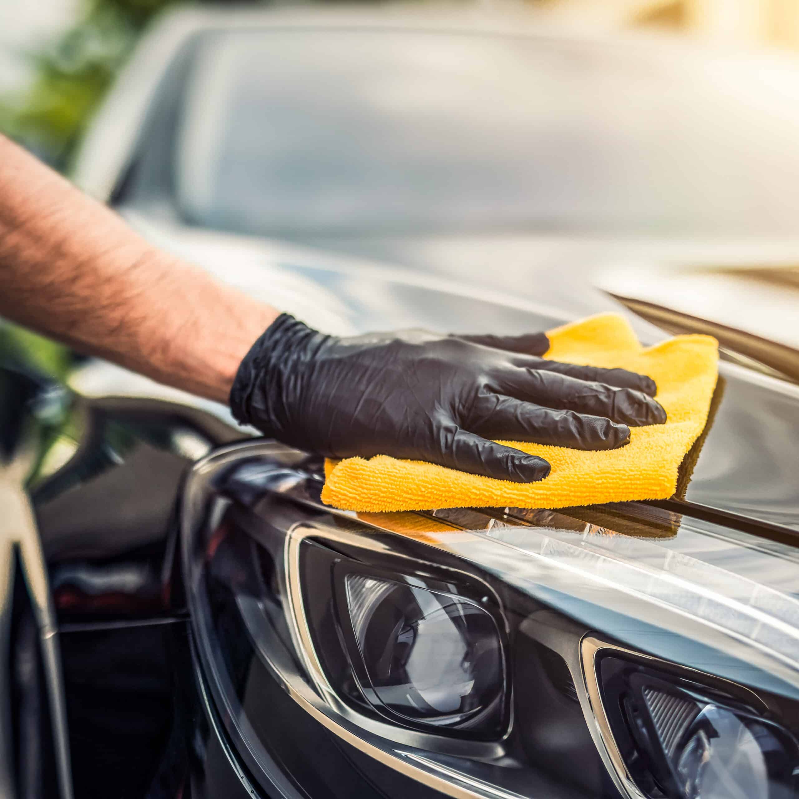 Main gantée de noir essuyant la peinture noire brillante d'une voiture avec une microfibre jaune près du phare.