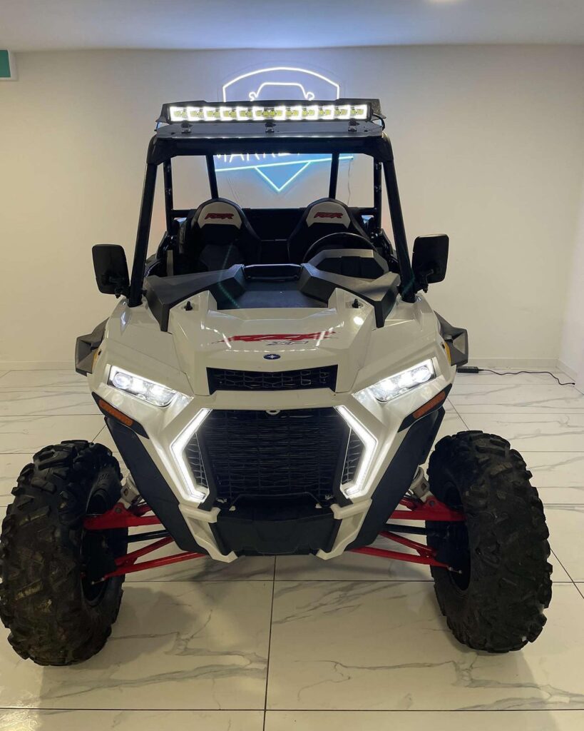 Polaris RZR Blanc Neuf : Aventure Tout-Terrain Polaris RZR blanc, neuf, avec phares LED allumés, barre lumineuse de toit, et suspension rouge dans un showroom.
