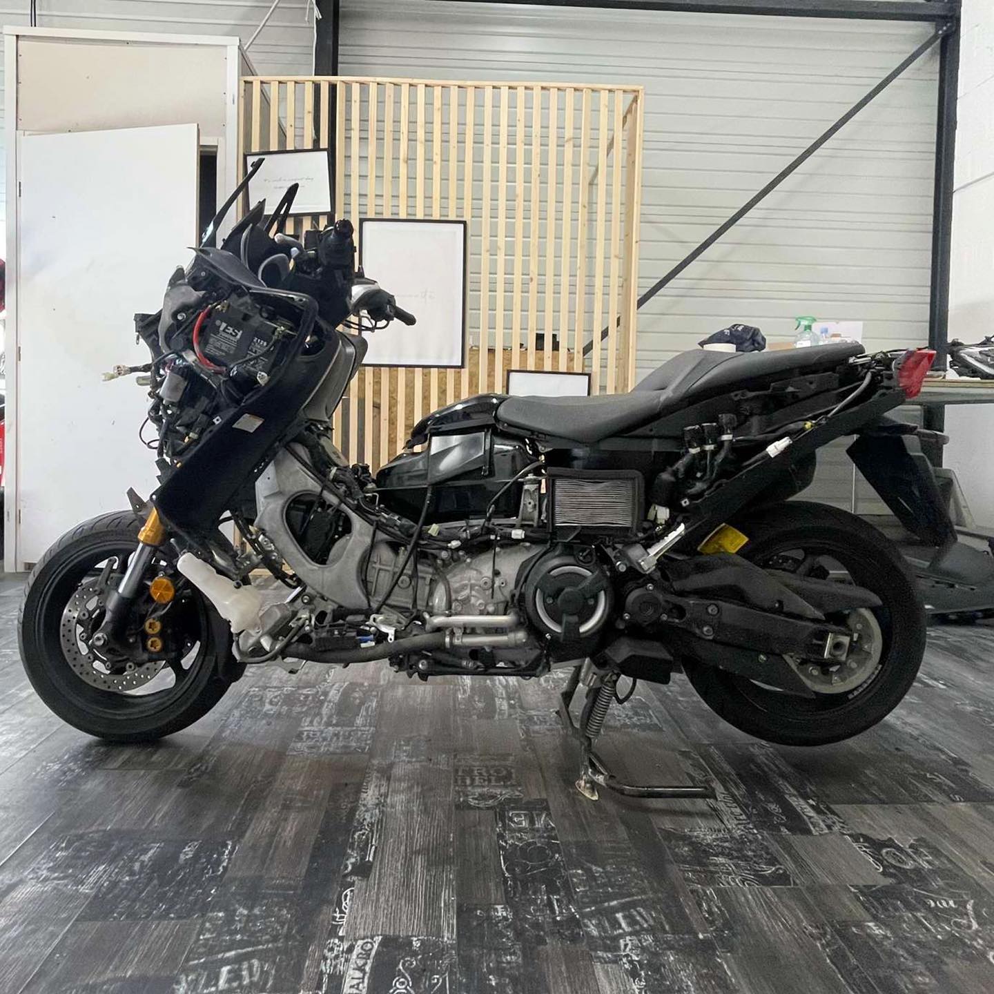 Maxi-scooter démonté, carénages retirés, exposant le moteur et la batterie avec l'inscription 'TES 12V' sur fond d'atelier.